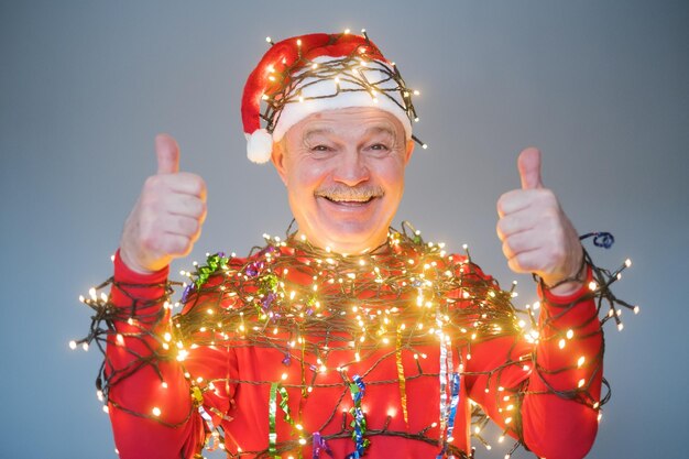 Senior man in rode kerstmuts gewikkeld in guirlande duim opdagen. Nieuwjaarsconcept 2023