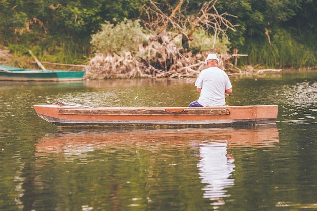 Senior man is fishing from a wooden boat in the center of a freshwater a lake fringed with lush scenery.