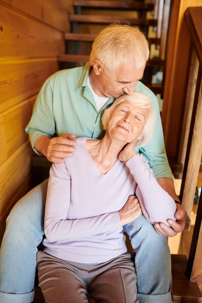 Photo senior man kissing his wife head and embracing her while both sitting on wooden staircase at home