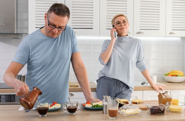 Senior man koken ontbijt en koffie voor zijn vrouw terwijl vrouw praten over de telefoon