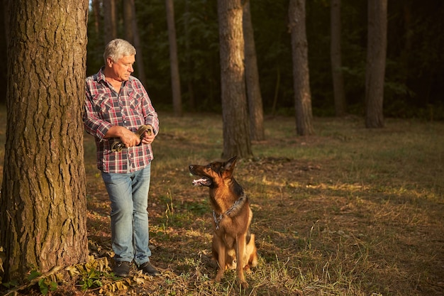 Senior man leaning on a tree while holding a leash and looking at his german shepherd pup