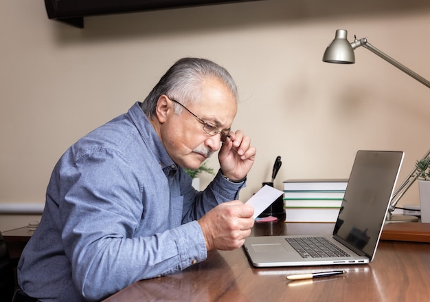 Senior man learn to use computer. Old man in glass and blue shirt Reads instructions for work using a laptop computer for online studying at home office