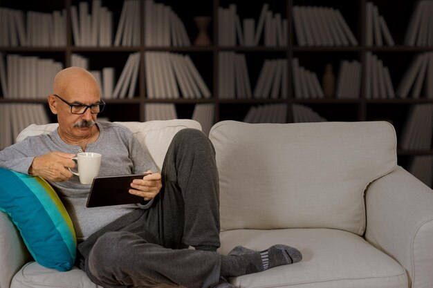 Senior man leest in een digitale tablet zittend op de bank in de huiskamer