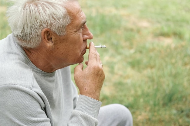 緑の自然を背景に屋外でタバコに火をつける年配の男性 思慮深い老人喫煙脇を見て顔を閉じるリラックスした年金受給者は外で週末の休息を楽しむ不健康な習慣