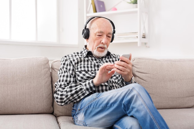 Senior man listening to music on smartphone with headphones, sitting on sofa at home, copy space