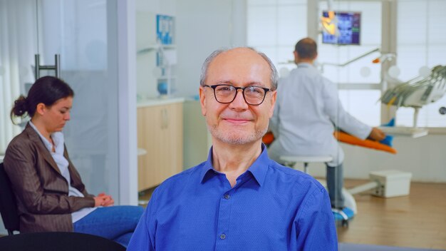 Senior man looking at camera while doctor examining patient in\
background. elderly male smiling on webcam sitting on chair in\
waiting room of stomatological clinic, assistant typing on pc