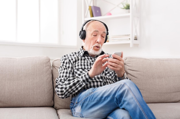 Senior man luisteren naar muziek op smartphone met koptelefoon, zittend op de bank thuis, kopieer ruimte