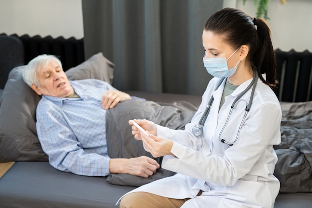 Senior man lying on couch next to young female doctor in whitecoat looking at thermometer after measuring his body temperature