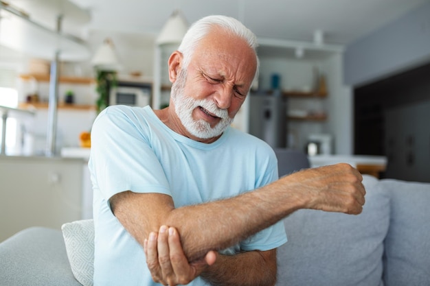 Senior man met armpijn Oude man die pijnlijke hand binnenshuis masseert Oude man hand met zijn elleboog die lijdt aan elleboogpijn Senior man die lijdt aan pijn in de hand thuis Ouderdom gezondheid