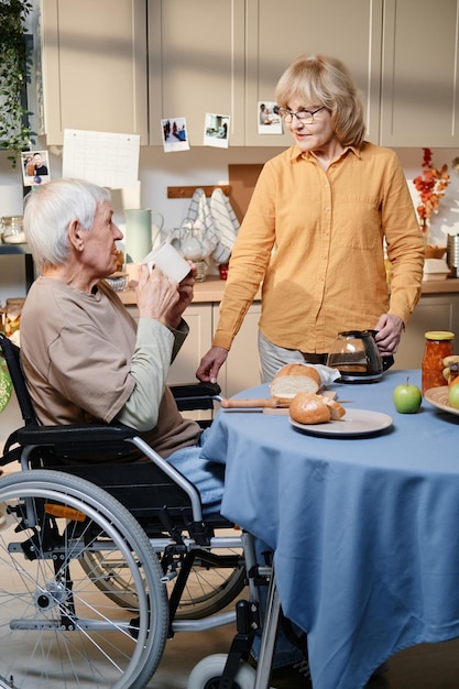 Senior man met rolstoel die thee drinkt en met zijn vrouw praat terwijl ze ontbijt kookt in de keuken