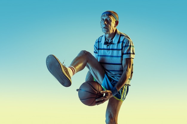 Senior man met sportkleding die basketbal speelt op gradiëntachtergrond, neonlicht. Kaukasisch mannelijk model in topvorm blijft actief. Concept van sport, activiteit, beweging, welzijn, vertrouwen.