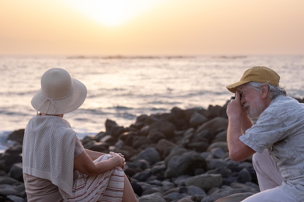 Senior man neemt een foto naar zijn vrouw die bij zonsondergang op het strand zit