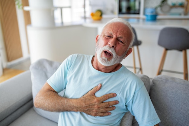 Senior man presses hand to chest has heart attack suffers from unbearable pain Mature man with pain on heart in living room Senior man suffering from bad pain in his chest heart attack at home