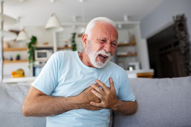 Senior man presses hand to chest has heart attack suffers from unbearable pain Mature man with pain on heart in living room Senior man suffering from bad pain in his chest heart attack at home