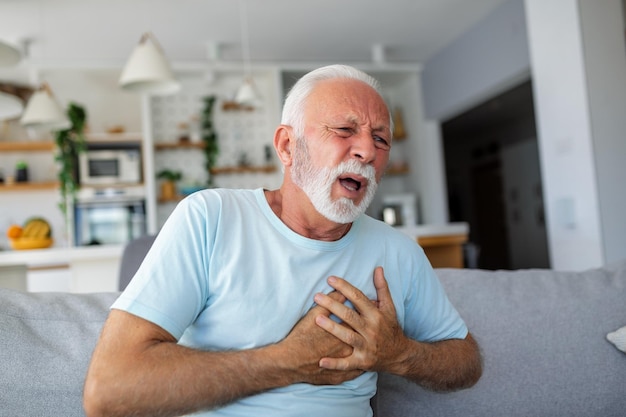 写真 年配の男性が手を胸に押し付け、心臓発作が耐え難い痛みに苦しんでいる リビングルームで心臓に痛みを感じている成熟した男性