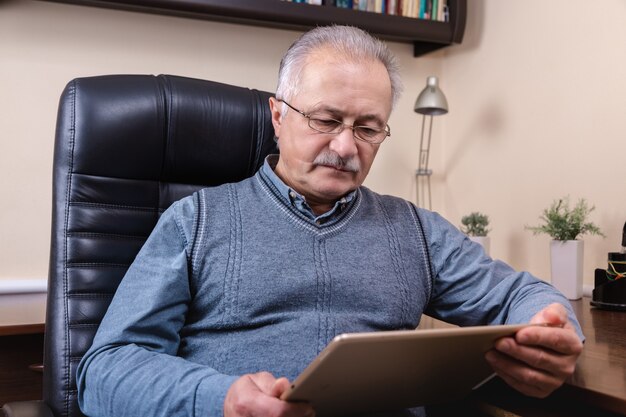 デジタルタブレットでニュースを読んでいる年配の男性。タブレットを使用して、自宅の机に座っている年配の男性。現代の技術、コミュニケーションの概念