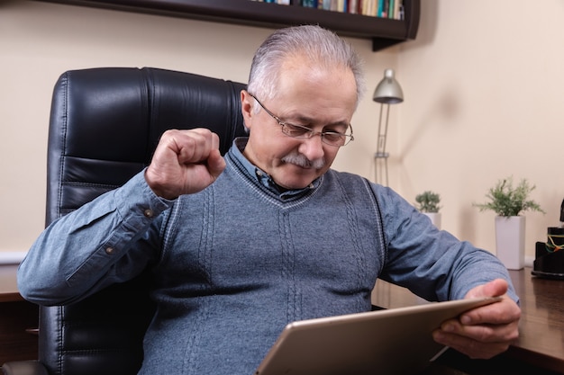 デジタルタブレットでニュースを読んでいる年配の男性。タブレットを使用して、自宅の机に座っている年配の男性。現代の技術、コミュニケーションの概念