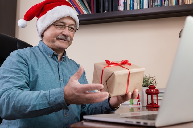 Senior man in santa claus hat Gives a gift and talks using laptop device for video call friends and childrens.