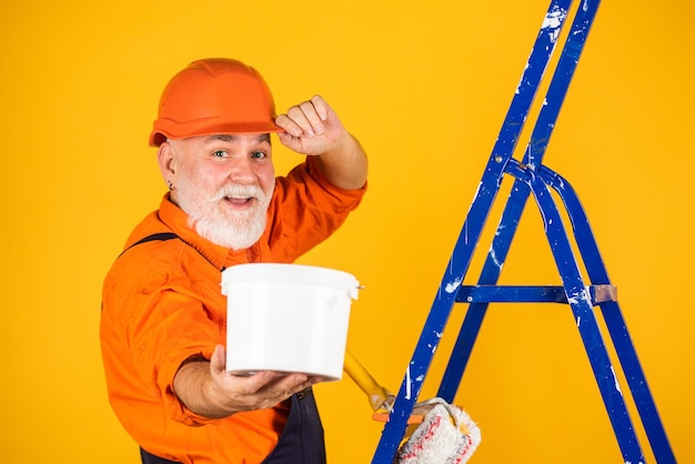 Senior man schilder gebruik roller op ladder. de muur geel schilderen. professionele schilder in werkkleding. werknemer schilderij muur in de kamer. mannelijke decorateur schilderen met roller. selectieve aandacht.