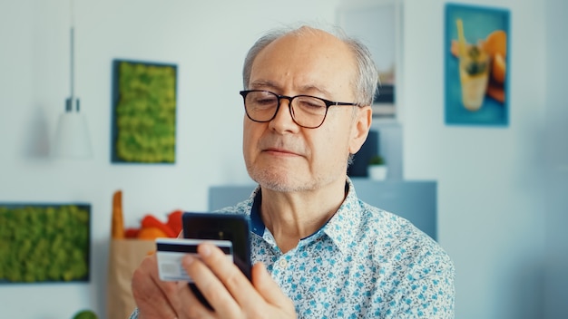 Senior man shopping online using smartphone and holding credit card during breakfast in kitchen. Retired elderly person using internet payment home bank buying with modern technology