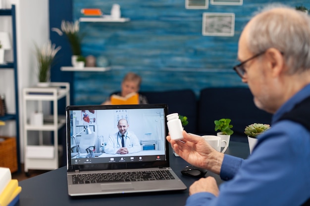 Uomo anziano che mostra la bottiglia di pillole del medico alla webcam durante la videochiamata uomo anziano che discute con la salute...