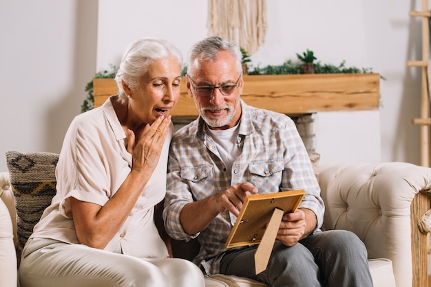 Photo senior man showing photo frame to her surprised wife sitting on sofa