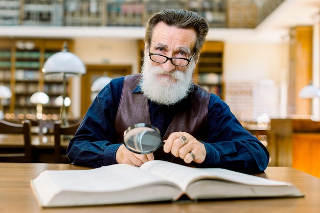 senior man sits in vintage library, holds magnifying glass and reads book. Bearded man in vintage shirt and leather vest working in library