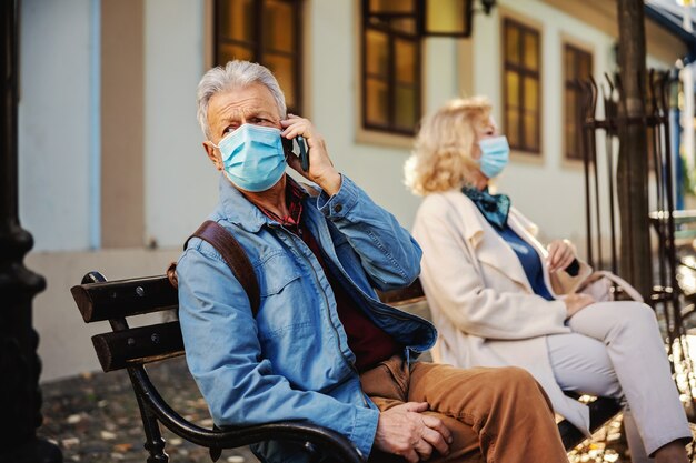 Senior man sitting on the bench outside. He is wearing protective face mask on.