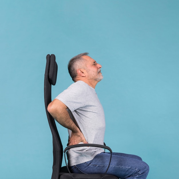 Uomo senior che si siede in sedia che ha dolore alla schiena su fondo blu