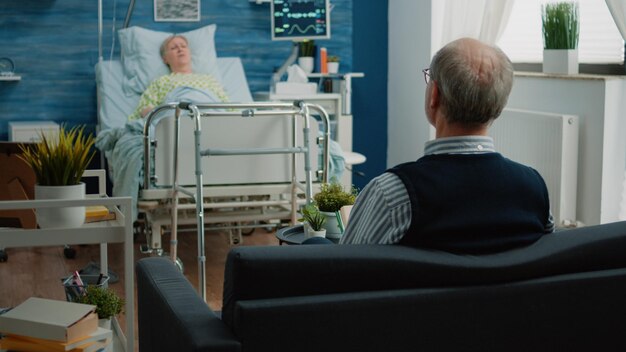 Senior man sitting on couch and talking to sick wife in hospital bed