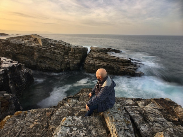 사진 해가 지는 동안 하늘을 배경으로 바다 의 바위 위에 앉아 있는 노인