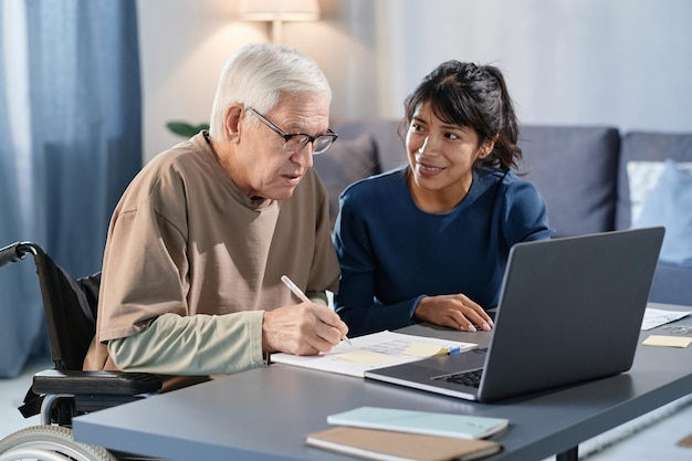 Senior man sitting at the table at home and filling documents with the help of volunteer