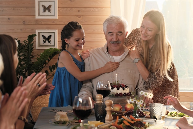 Senior man sitting at the table with birthday cake while his granddaughters congratulating him with birthday at home