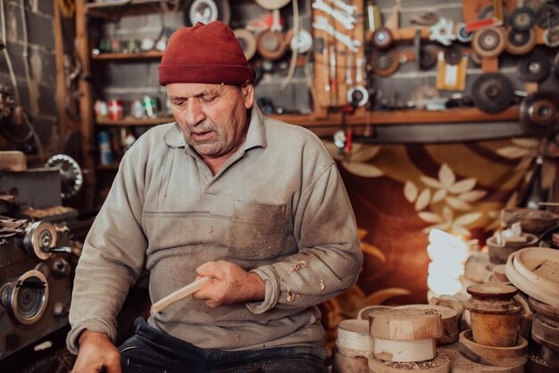 Старший мужчина сидит в мастерской и обрабатывает деревянную посуду старым ручным способом.