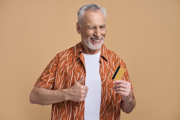 Senior man smiles winks shows thumb up at camera posing with bank credit card isolated on beige