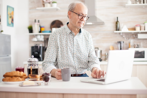 Senior man smiling and using laptop in the kitchen. Daily life of senior man in kitchen during breakfast using laptop holding a cup of coffee. Elderly retired person working from home, telecommuting u