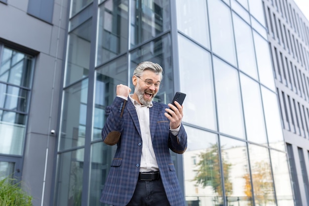 A senior man in a suit stands near an office center and is happy shows a victory gesture with his