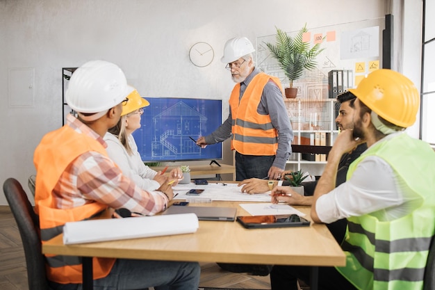 Senior man in uniform and helmet presenting new construction project to colleagues on tv screen