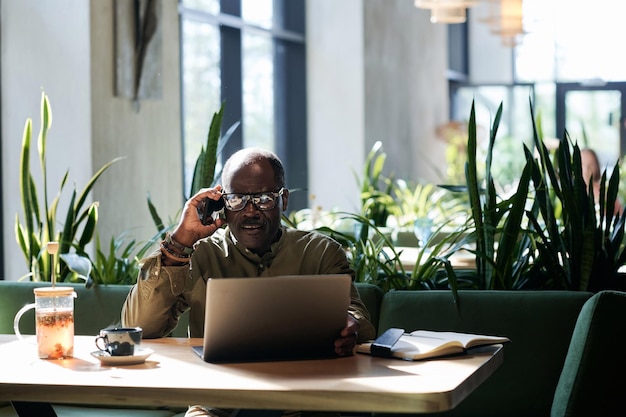 Senior man using laptop and talking on the phone while sitting in cafe