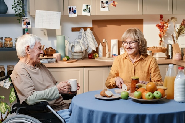 車椅子を使って妻と話している年配の男性、彼女は笑顔で台所のテーブルでパンを切っています