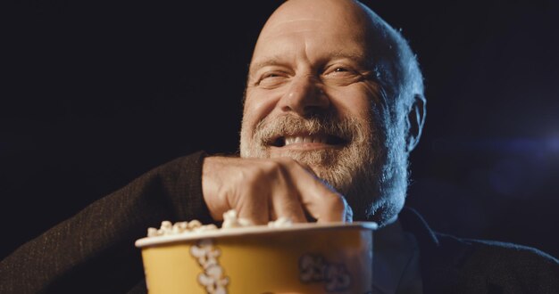 Старший мужчина смотрит смешной комедийный фильм в кинотеатре он смеется и ест попкорн