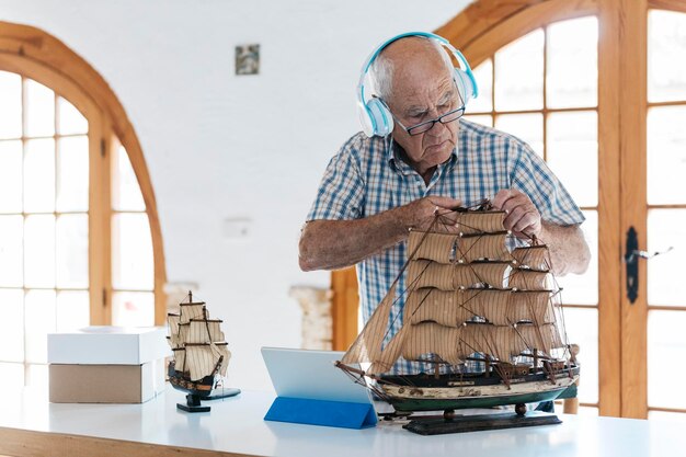 Senior man wearing headphones working on model ship on table with tablet