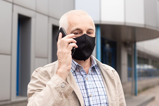 Senior man wearing mask using smartphone in city near office buildings