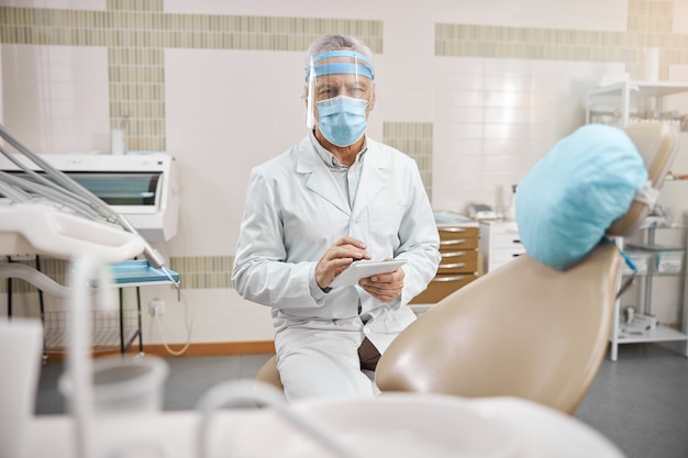 senior man wearing a surgical mask and holding a tablet while looking at the camera