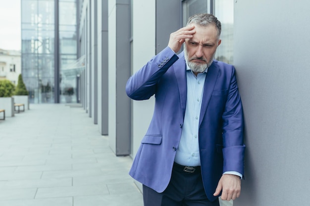 Senior man werknemer zakenman in een pak staat in de buurt van het kantoorcentrum leunt tegen de muur