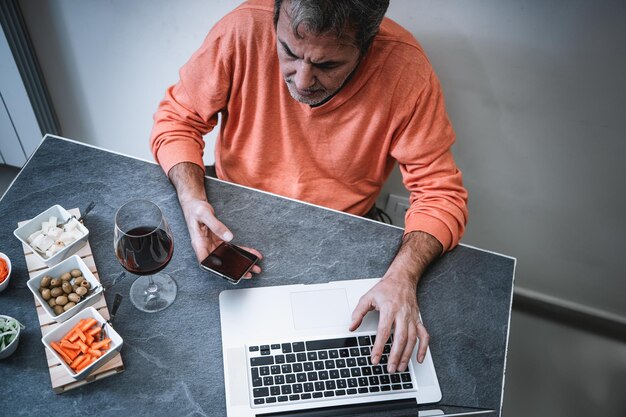 Senior man werkt op afstand vanuit huis zittend in de keuken met een laptopcomputer