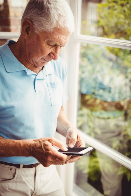 Старший мужчина рядом с окном с помощью смартфона на дому