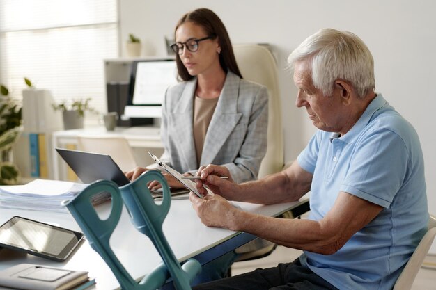 Photo senior man with disability signing document during meeting with social worker