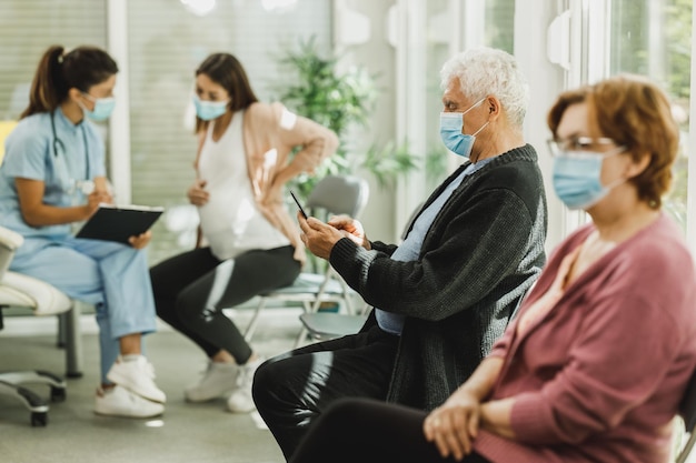 Senior man with a face mask sitting in a waiting room of a hospital and surfing at his smartphone.