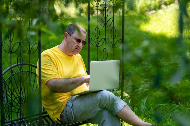 Старший мужчина с ноутбуком, работающий снаружи в концепции домашнего офиса в зеленом саду